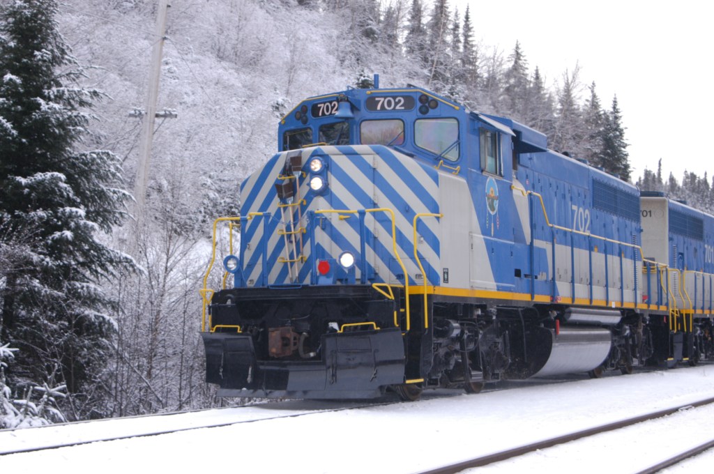 Blue and grey train engine on a snow covered track flanked by spruce trees.